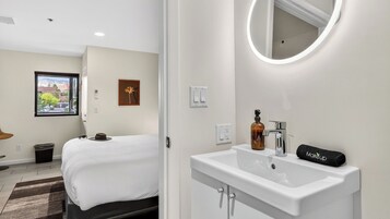 A hotel room with a bed, a bathroom with a sink and mirror, a window with a view, and a wall-mounted light fixture.