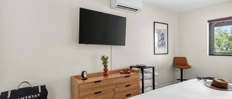 A modern hotel room with a flat-screen TV, a bedside table with a vase and book, a wooden dresser, a chair, and a window with a view.