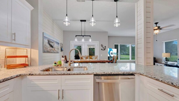 Una cocina moderna con armarios blancos, encimeras de granito, lavavajillas de acero inoxidable, iluminación colgante y vistas al exterior.