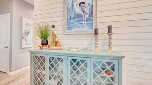 Un mueble azul claro con puertas de cristal, un cuadro enmarco de un pájaro, una planta en maceta y velas.
