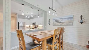 Un comedor con mesa y sillas de madera, iluminación colgante, una cocina al fondo y un cuadro en la pared.