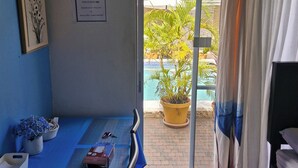 Een kamer met een bed, een bureau met een computer, een stoel, een kamerplant en uitzicht op een buitenzwembad.