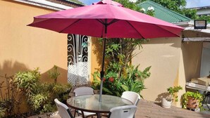 Een terras met een glazen tafel, witte stoelen, een rode parasol en potplanten.