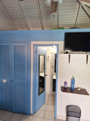 A room with a closed blue door, a white sink, a mirror, a television, and a water bottle on a small table. - Cozy, convenient and secured studio-Located Almond Grove Cole bay. (Cole Bay)