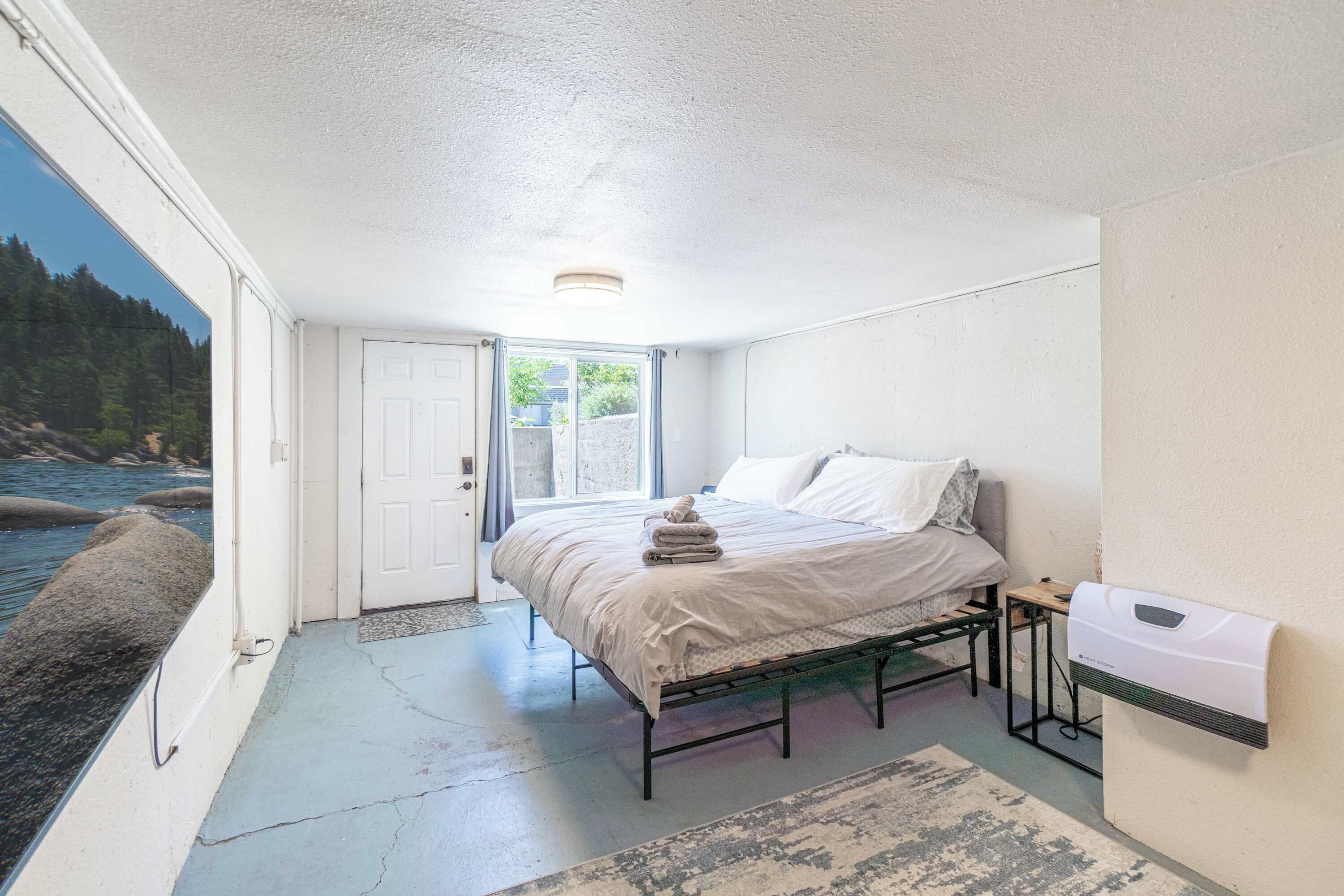 A bedroom with a bed, a bedside table, a wall-mounted heater, and a view of the outdoors.