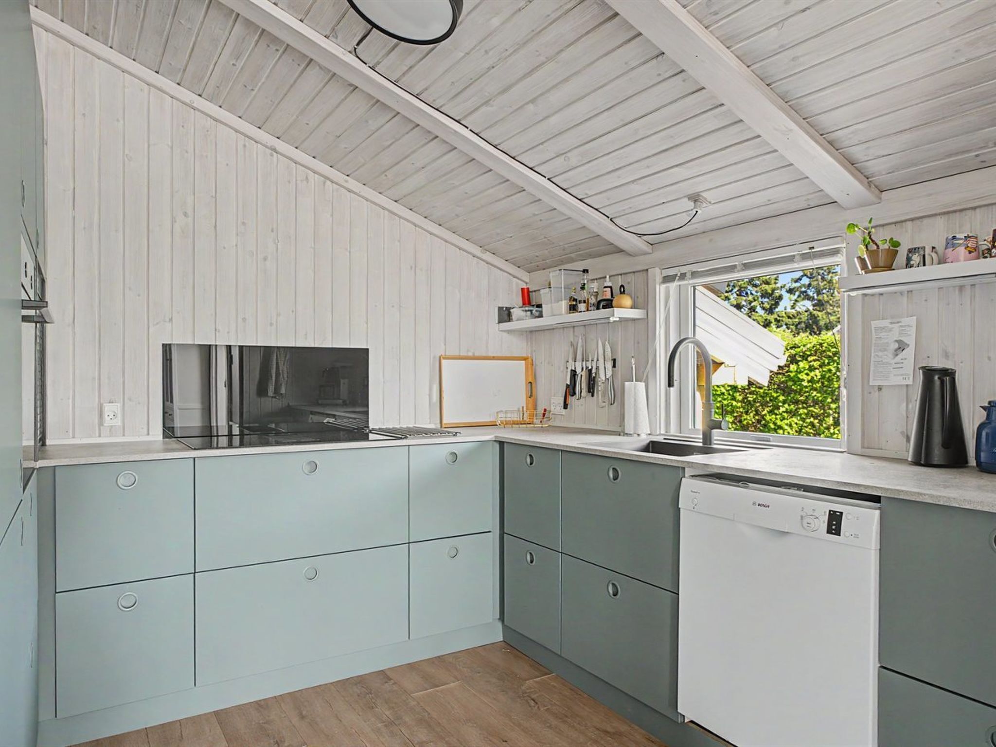 A kitchen with light blue cabinets, a black stove, a white dishwasher, and a window with a view of greenery.
