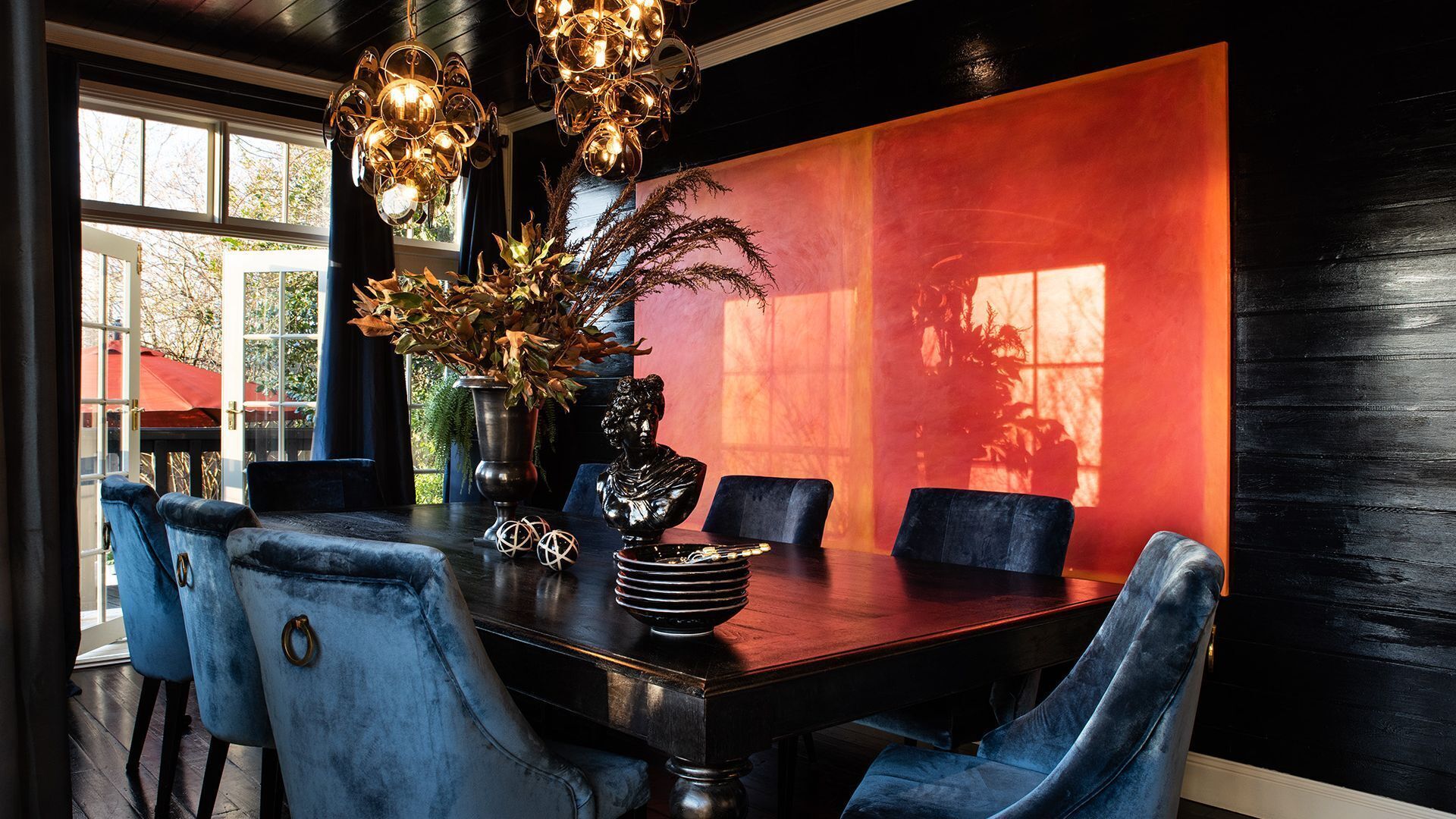 A dining area with a dark wooden table, blue upholstered chairs, a vase with dried plants, and a wall with a red and black gradient.