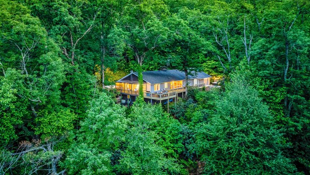 Uma casa rodeada por uma floresta densa, com um alpendre visível e várias janelas.