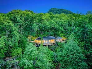 Une maison entourée d’une forêt dense, dotée d’une terrasse bien visible et de nombreuses fenêtres.