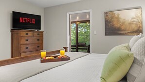 Quarto com cama, TV exibindo 'NETFLIX', cômoda de madeira, bandeja com café da manhã e janela com um letreiro 'WELCOME'.