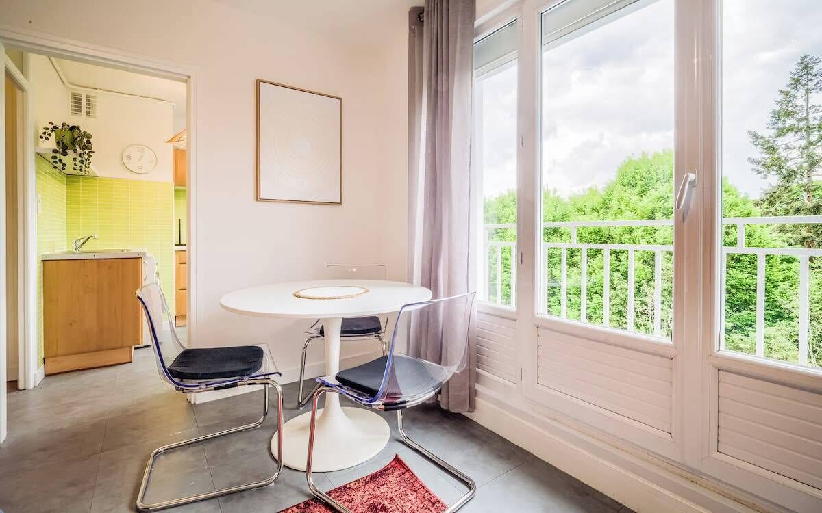 A room with a dining table, chairs, a view of greenery outside, and a kitchen area with a sink and cabinets.