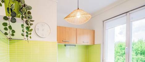 A kitchen with a sink, wooden cabinets, a white countertop, a green-tiled backsplash, a window, and a hanging plant.