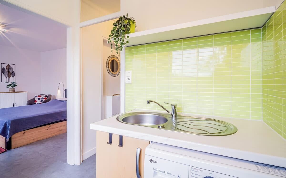 Una cocina compacta con lavamanos, lavadora y una cama en la habitación contigua.
