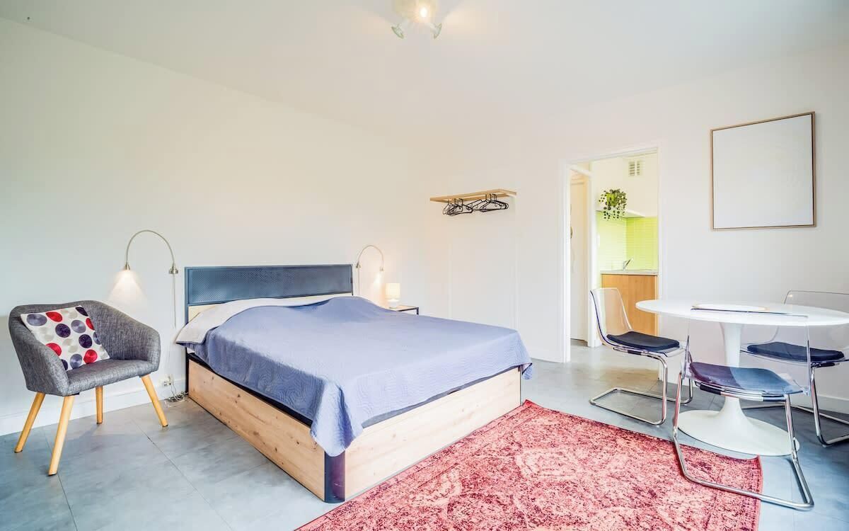 A modern bedroom with a bed, bedside lamps, a chair, a table, and a view of a kitchen.