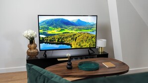 Uma televisão de tela plana sobre um suporte preto, um vaso com flores brancas, um tabuleiro verde-água e um caderno sobre uma mesa de madeira.