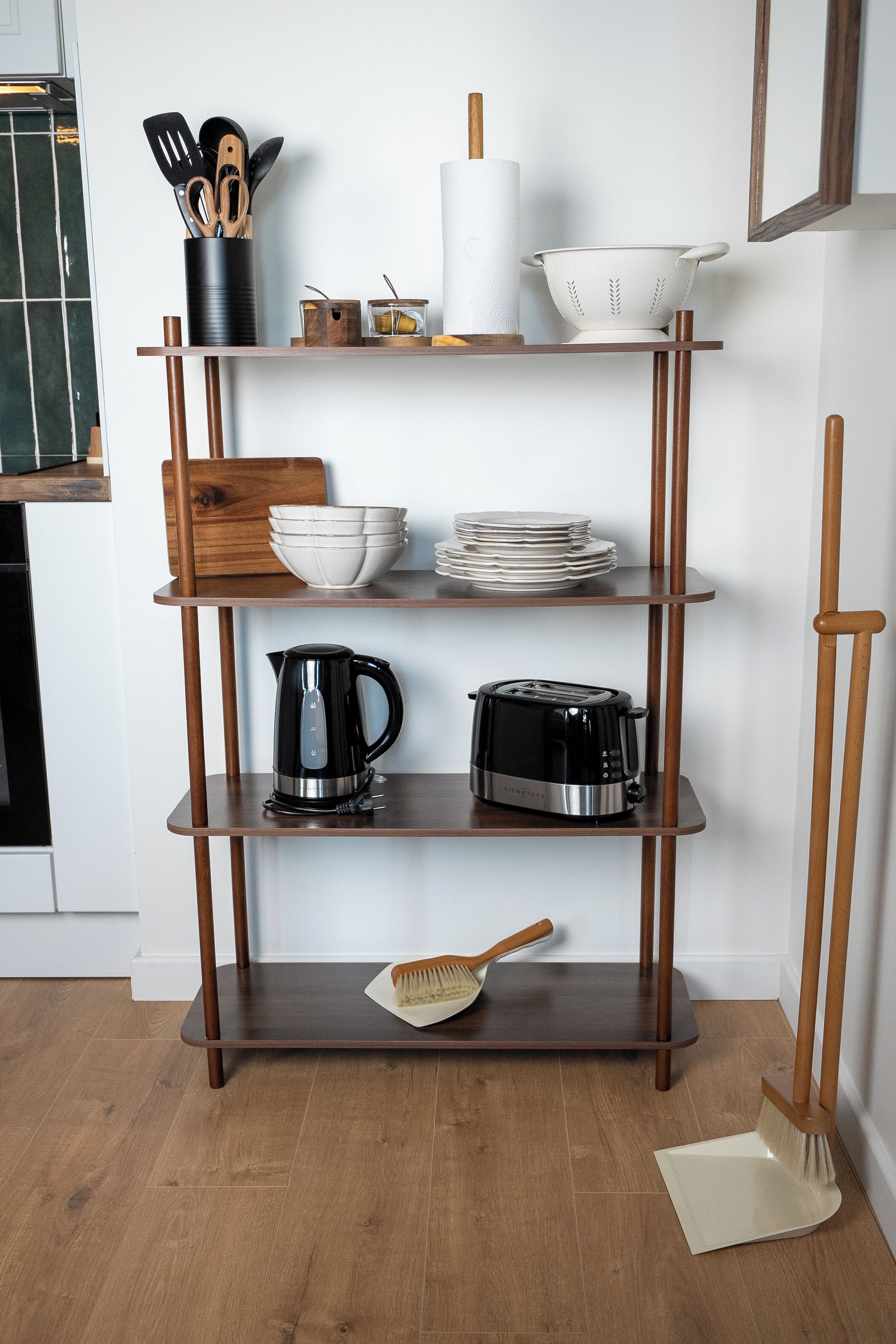 Una cocina con una estantería de madera que contiene utensilios de cocina, un hervidor, un tostador y una escoba.