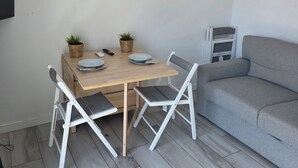 Ett rum med ett matbord i trä, två vita stolar, en grå soffa och en liten tv på väggen.