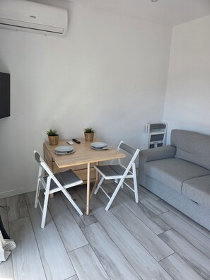 Une pièce comprenant une table à manger en bois, deux chaises blanches, un canapé gris et un petit téléviseur fixé au mur.