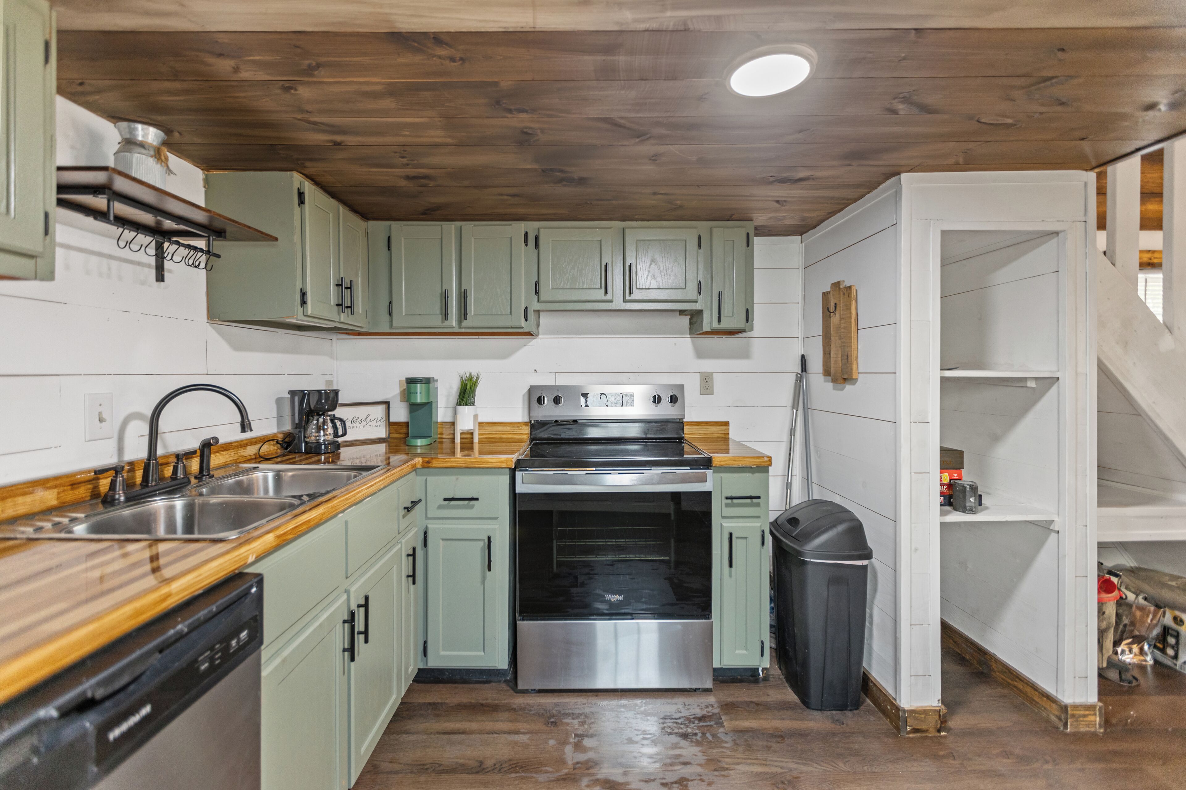 Una cocina con gabinetes verdes, una estufa de acero inoxidable, un lavavajillas y un cubo de basura.