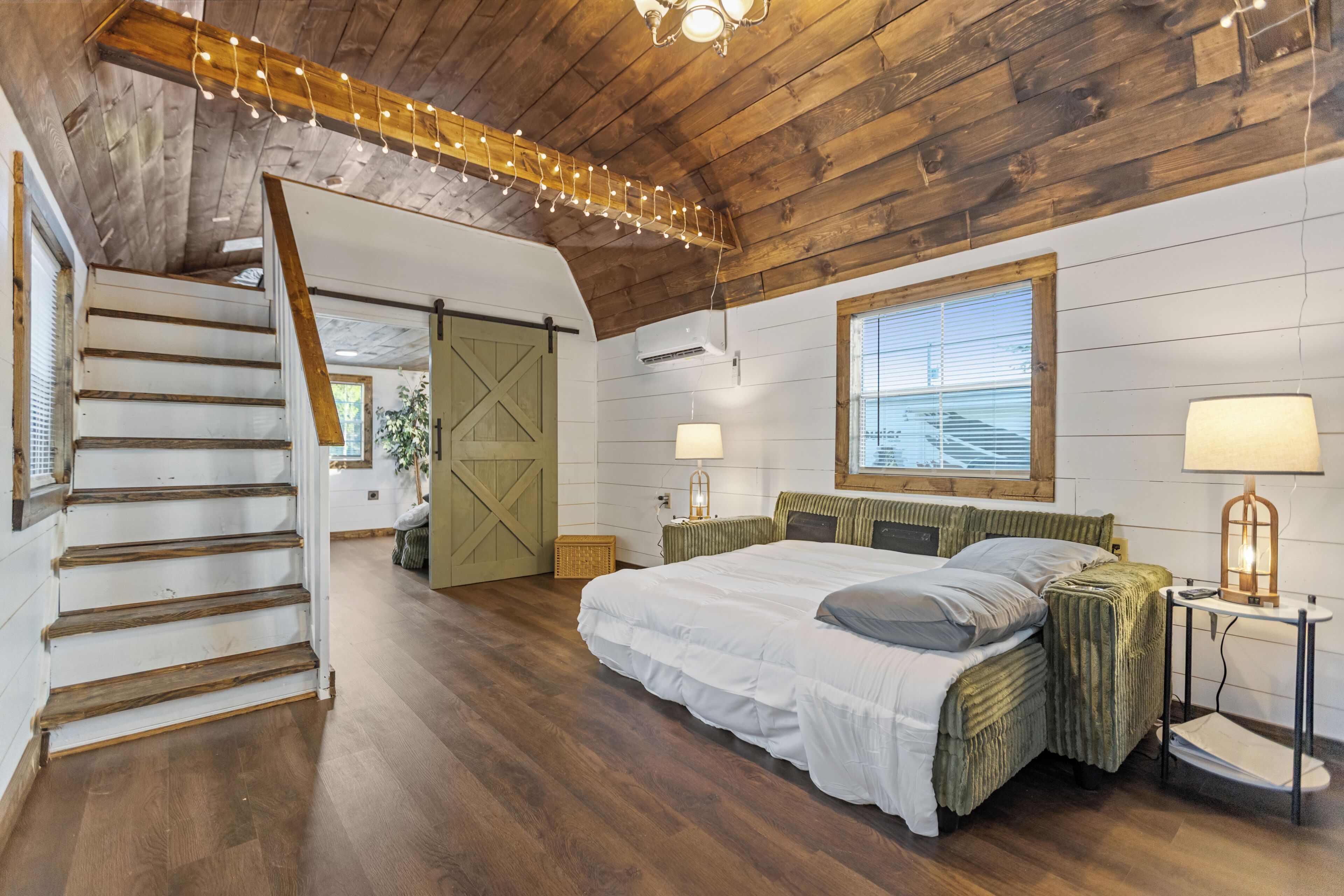 Une chambre avec un escalier en bois, un lit doté d’une tête de lit verte, une fenêtre avec des stores et une lampe sur une table de chevet.