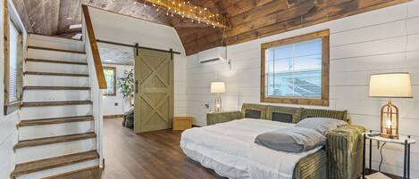 Une chambre avec un escalier en bois, un lit doté d’une tête de lit verte, une fenêtre avec des stores et une lampe sur une table de chevet.