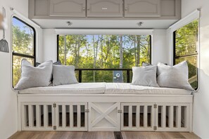 A white bench with cushioned seating and storage cabinets underneath, positioned in front of a window with a view of trees. - New Tiny Home + Camper - Sleeps up to 10 - Fire Pits - Access to Brimstone Trail (Huntsville)