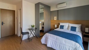Quarto de hotel com cama, escrivaninha, cadeira, mesinha com planta e espelho.