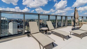 Um lounge no terraço com espreguiçadeiras, uma mesa pequena e um guarda-sol, com vista para uma paisagem urbana com edifícios e céu claro.