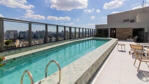 Piscina no terraço com vista para a cidade, guarda-corpo de vidro e área de estar externa.