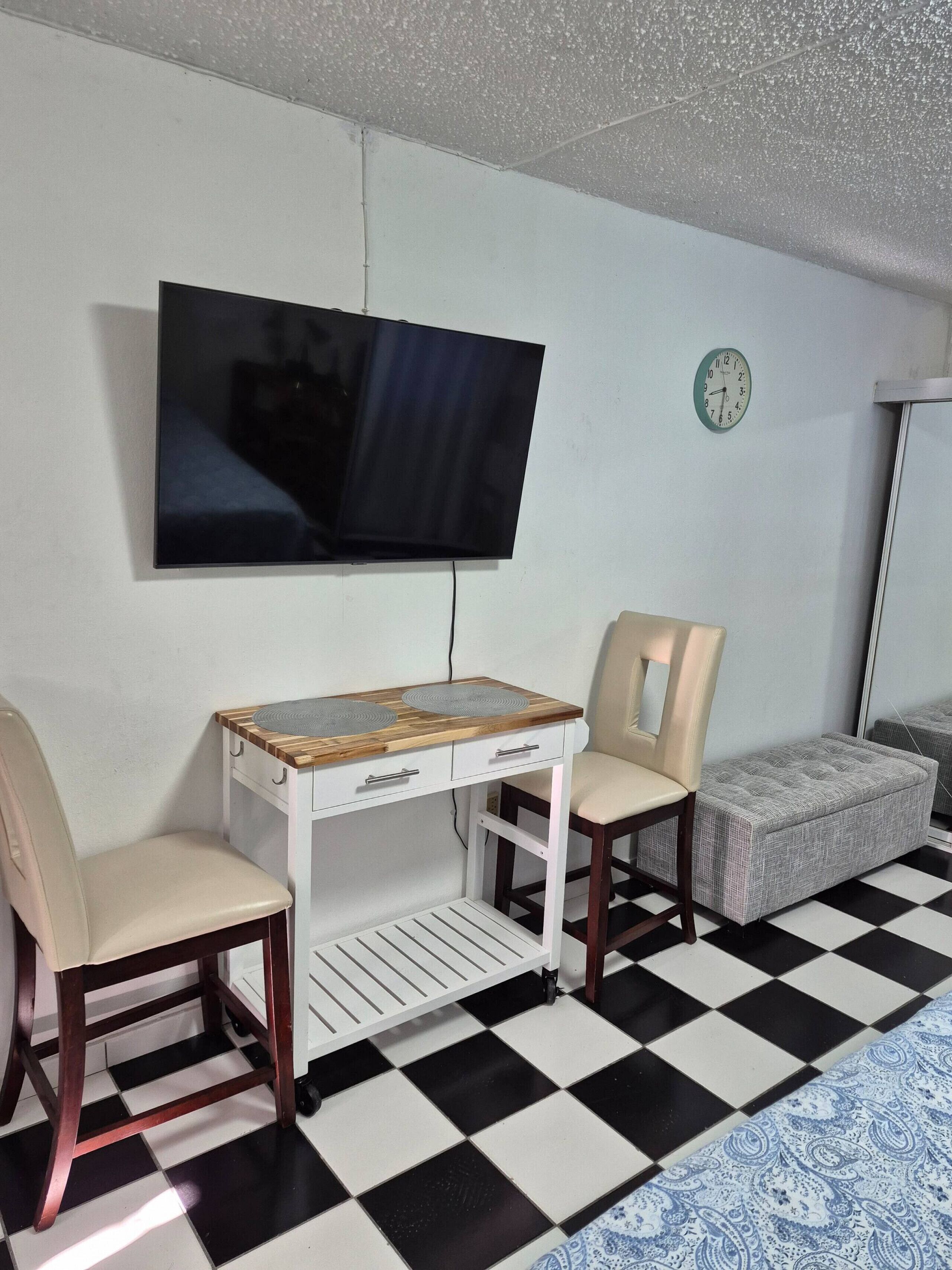 A room with a TV mounted on the wall, a small white table with drawers, two chairs, a clock, and a patterned rug.
