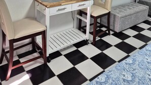 Una stanza con una TV fissata al muro, un piccolo tavolo bianco con cassetti, due sedie, una sveglia e un tappeto decorato.