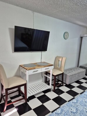 Ein Zimmer mit an der Wand montiertem Fernseher, einem kleinen weißen Tisch mit Schubladen, zwei Stühlen, einer Uhr und einem gemusterten Teppich.