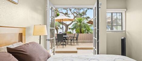 A hotel room with a bed, a desk, and an open door leading to an outdoor seating area with chairs and umbrellas.