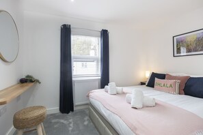 A hotel room with a bed, a bedside table, a chair, a window with curtains, a radiator, and a wall-mounted picture. - Stylish Two-Bed Cheltenham Retreat with Hot Tub (Cheltenham)