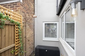 A narrow outdoor space with a wooden fence, a window with blinds, and a black heater.