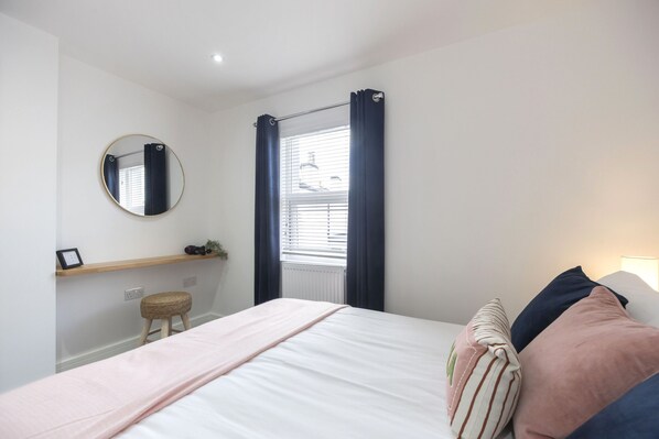 A bedroom with a bed, a round mirror, a window with curtains, a shelf with a picture frame, and a small stool.