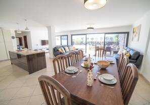 On aperçoit un coin repas avec une table en bois dressée pour quatre personnes, entourée de chaises. Au fond, on distingue une cuisine avec un réfrigérateur et un évier.