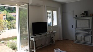 A room with a TV on a stand, a cabinet, a sliding glass door, and a view of greenery outside. - Thieman House - MENTON (MENTON)