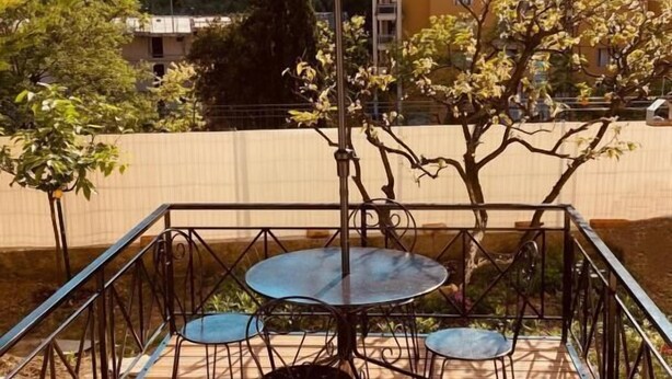 An outdoor patio with a table and chairs, a large umbrella, and a view of a hill with buildings. - Thieman House - MENTON (MENTON)