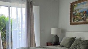 Ein Schlafzimmer mit Bett, Nachttisch, Lampe, einem Gemälde und Blick auf Grünflächen.