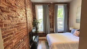A bedroom with a brick wall, a bed, a desk, a plant, and a ceiling fan. - Benton Park Walkable Apartment- IN1S (St. Louis)