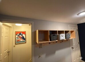 Un couloir orné d’une affiche sur le thème du ski, une étagère en bois avec des paniers de rangement et une porte blanche.