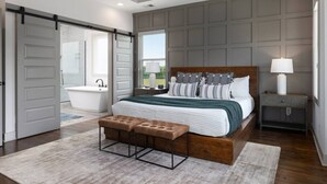 A bedroom with a large bed, a wooden bench, a nightstand, a lamp, and a view of a bathroom. - House with Rooftop Patio, Next to a Park, 3 Miles to Downtown (Nashville)