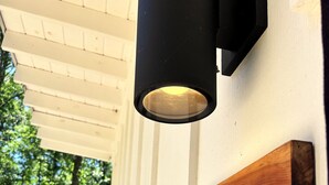 Une enseigne en bois portant le nom « Magnolia », fixée à un mur blanc, un luminaire extérieur noir placé au-dessus, et une vue sur une zone boisée à travers une fenêtre.