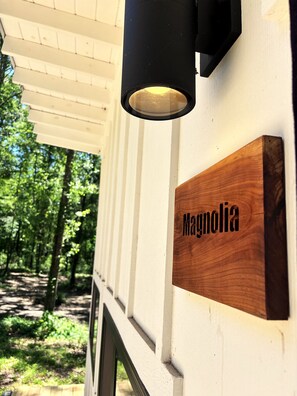 A wooden sign with the name 'Magnolia' mounted on a white wall, a black outdoor light fixture above it, and a view of a forested area through a window.