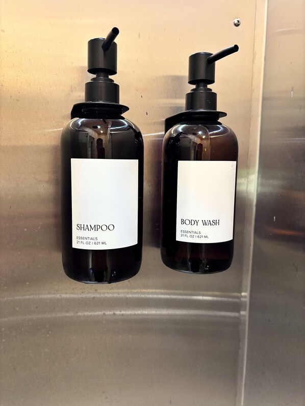 Two bottles of shampoo and body wash with pump dispensers, labeled 'SHAMPOO' and 'BODY WASH,' respectively, on a stainless steel surface.