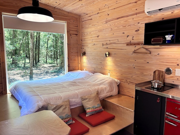 A wooden cabin interior with a bed, a small table, a red cabinet, and a window overlooking a forest.