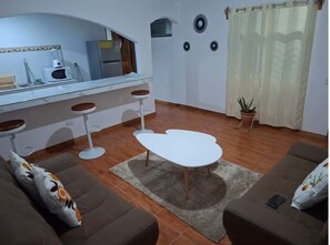 A living room with a sofa, a coffee table, a kitchen area with a microwave and refrigerator, and a potted plant. - Villa Ensueño Cahuita 2 familiar (Westfalia)