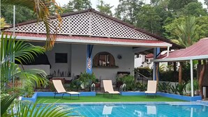 A swimming pool with lounge chairs, a covered patio, and a house with a lattice roof. - Alojamientos Ensueño Cahuita 1 (Westfalia)