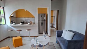 Un salon moderne comprenant un canapé, des poufs, une table basse et un coin cuisine avec un réfrigérateur et un plan de travail.